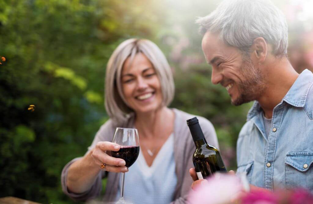 A couple smiling is pouring a bottle of red wine in glasses.